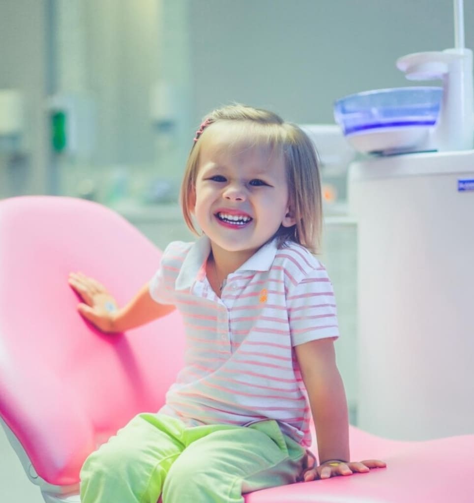nina sonriendo sentada en sillon dental en clinica con equipo medico al fondo