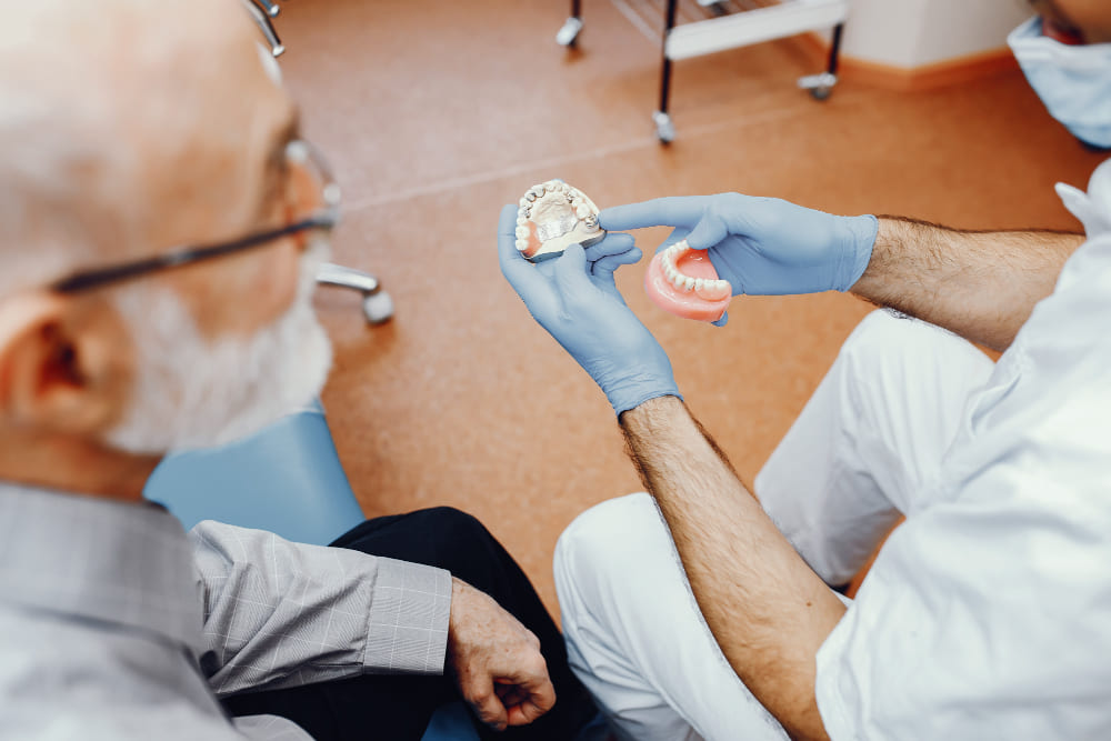 hombre mayor sentado en consulta dental observando protesis que sujeta dentista con guantes